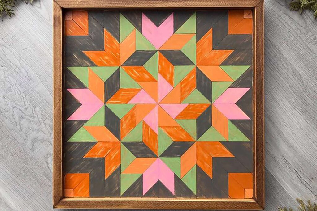 a framed barn quilt laying on the wooden floor. Orange, Green & Pink coloured