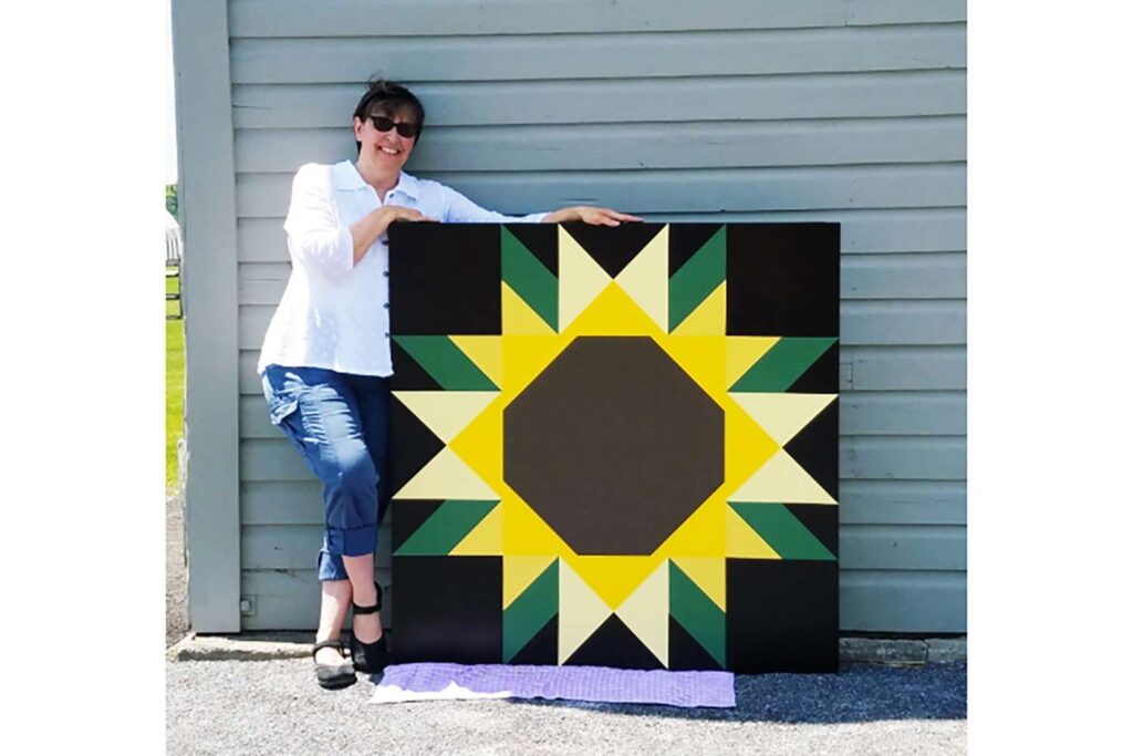 a quilter holds a sunflower pattern barn quilt outside her garage.
