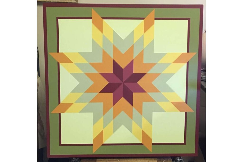 a barn quilt with a pattern of stars is placed on a wooden shelf.