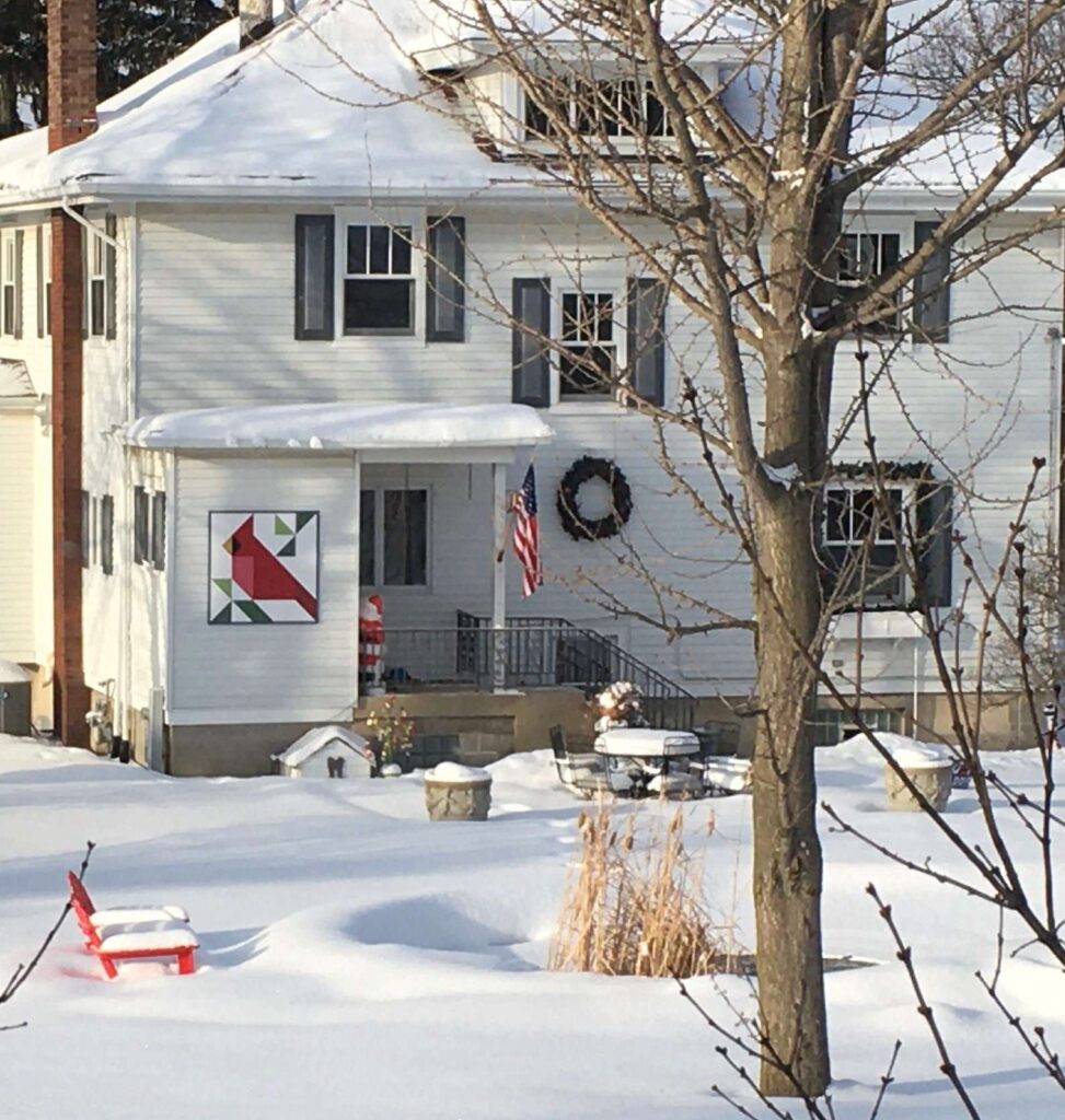 a barn quilt hanging on the a big house surrounded by white snow.