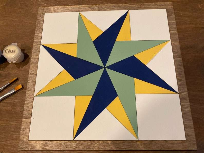 a pinwheel barn quilt laying on the wooden floor, next to a color box and two brush.