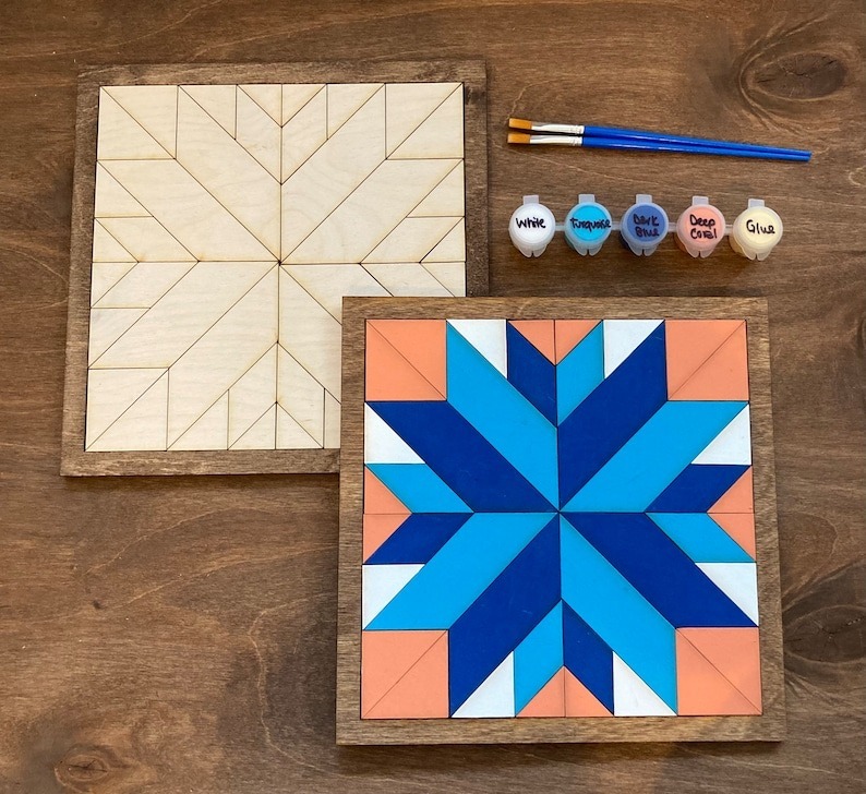 two small barn quilt laying on the wooden floor, next to 5 color boxes and a brush.