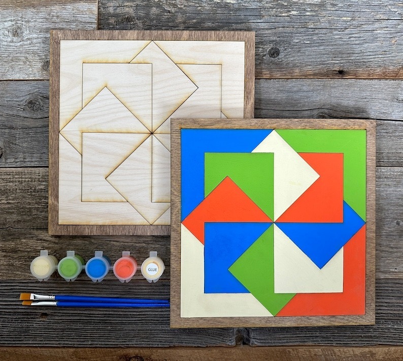 two barn quilt laying on the wooden floor, next to five color boxes and two brush.