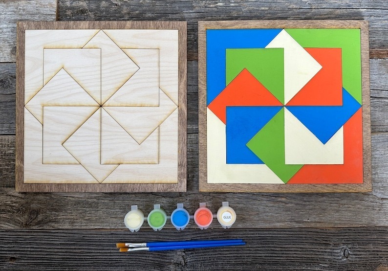 two barn quilt laying on the wooden floor, next to five color boxes and two brush.
