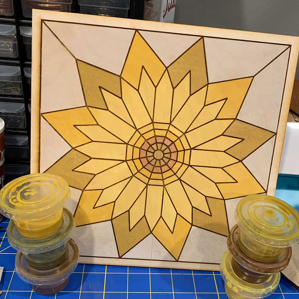 a barn quilt with sunflower pattern placed on the table, behind six color boxes