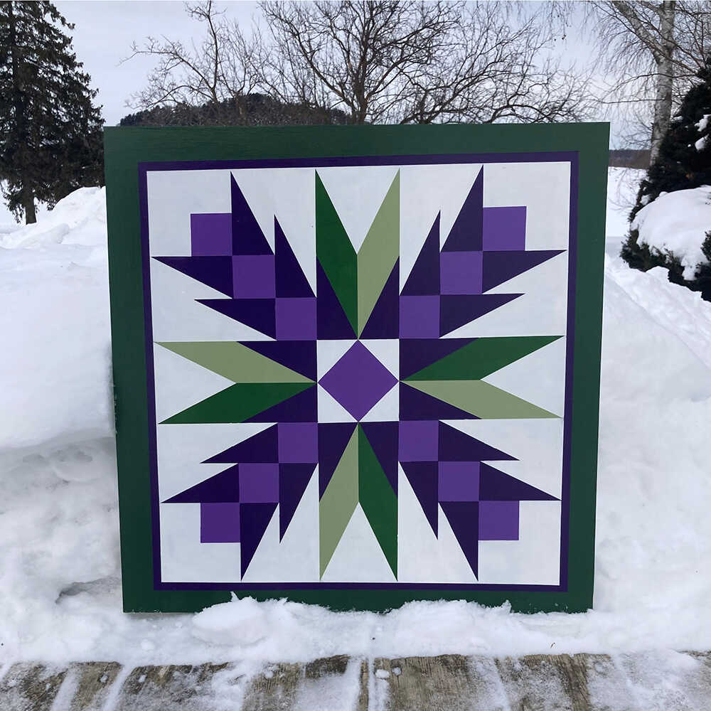 a barn quilt with purple flowers pattern placed on the snow in the forest.