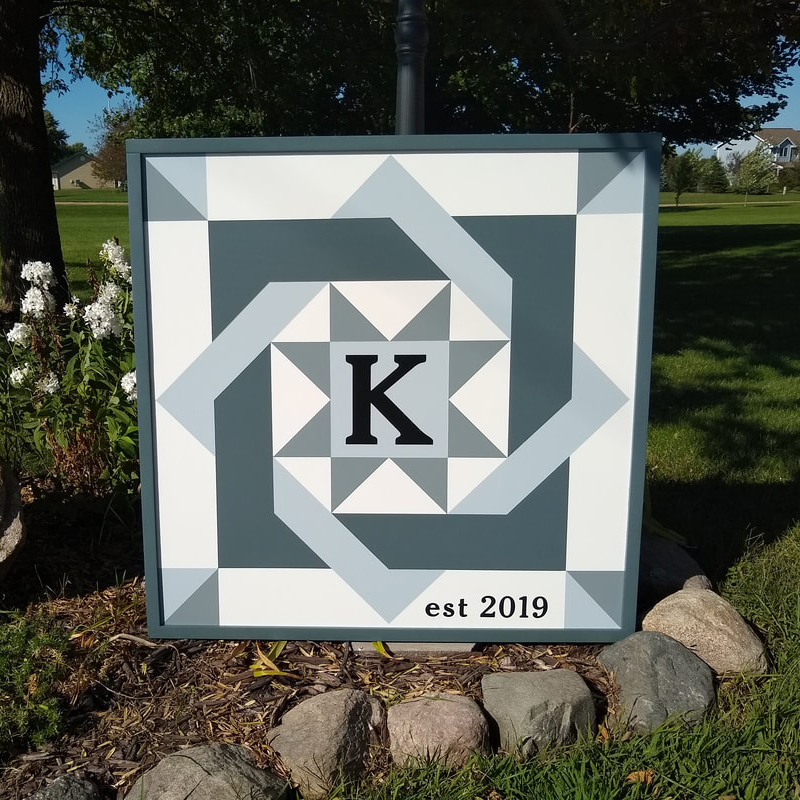 a gray barn quilt with an interlocking squares pattern leans against a black lamppost in the garden