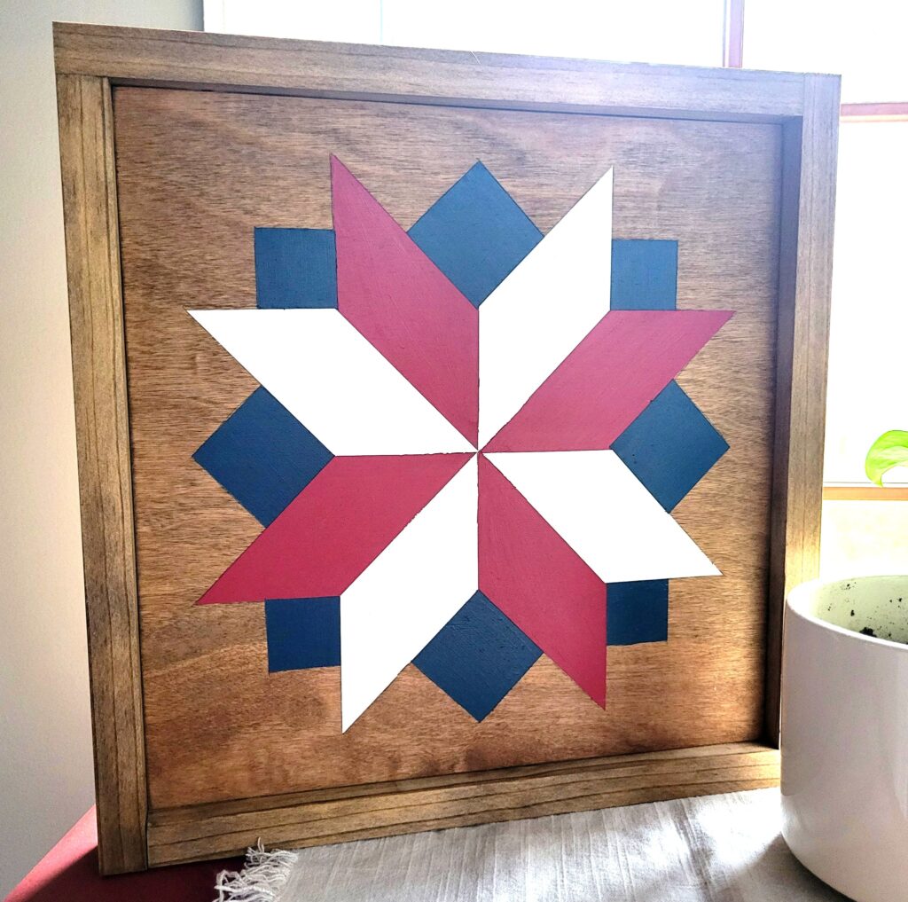 a barn quilt placed on the table, next to a potted plant.