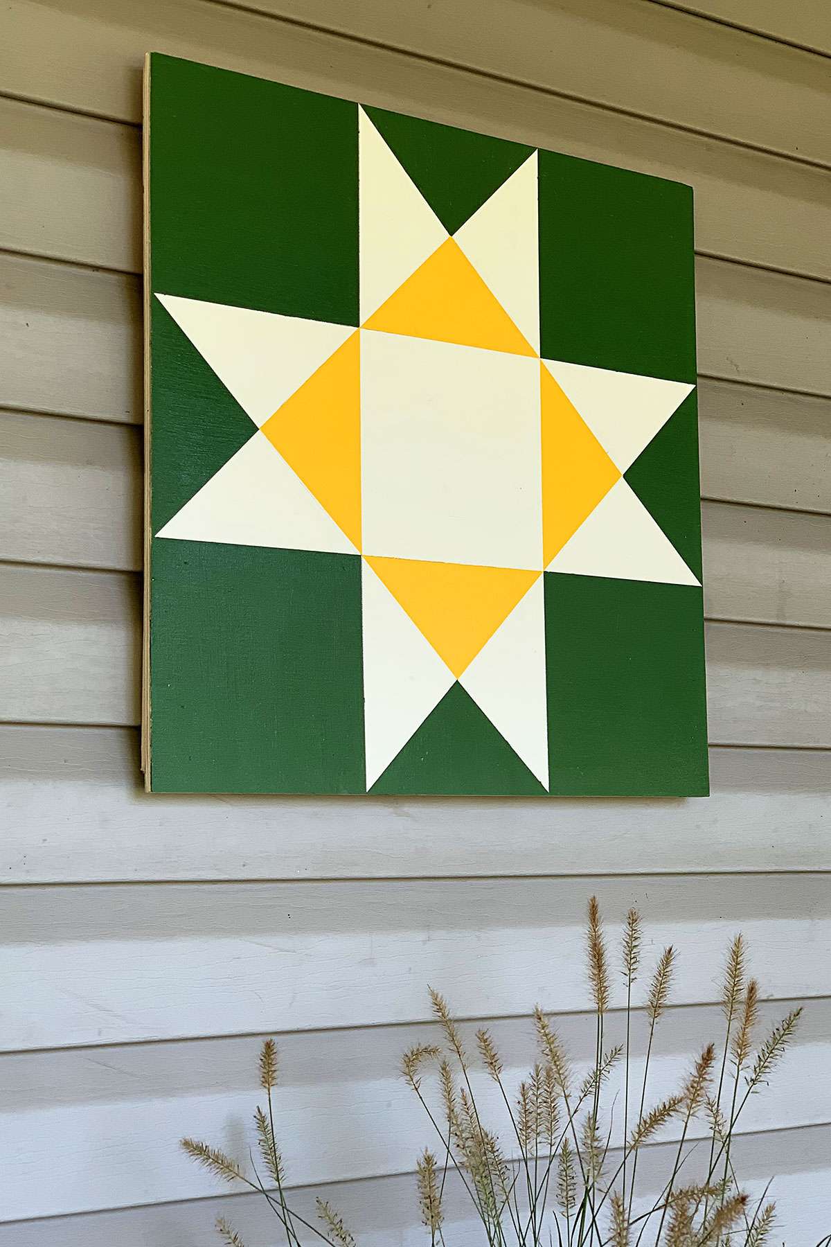 a white and yellow star barn quilt hanging in the wall, above a grass bush.
