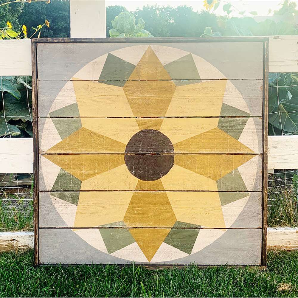 a sunflower barn quilt placed on the grass, leaning against the wooden fence