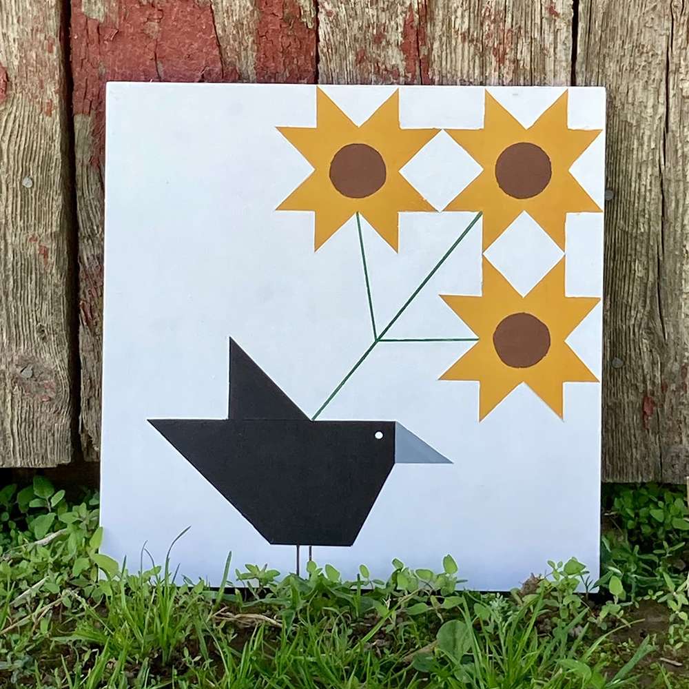 a barn quilt with the a black crow and three sunflowers placed on the grass