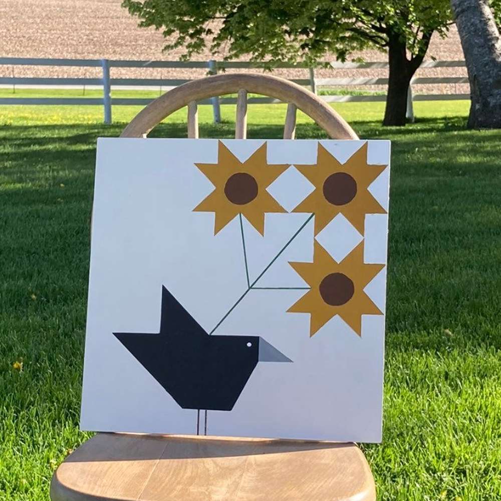 a barn quilt with the black crow and sunflower patterns placed on an outdoor wooden chair