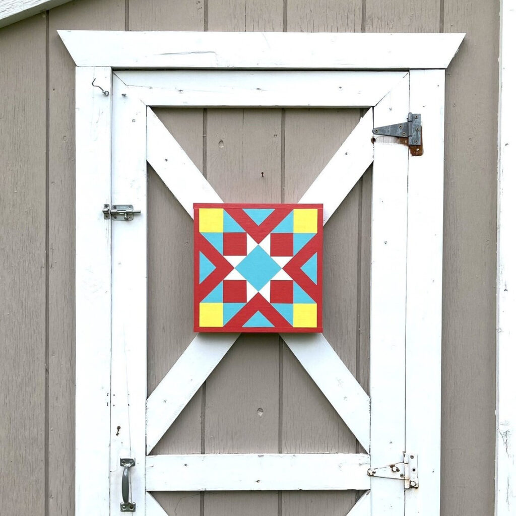 the barn quilt with red and yellow triangles and square patterns hanging on the wood window.