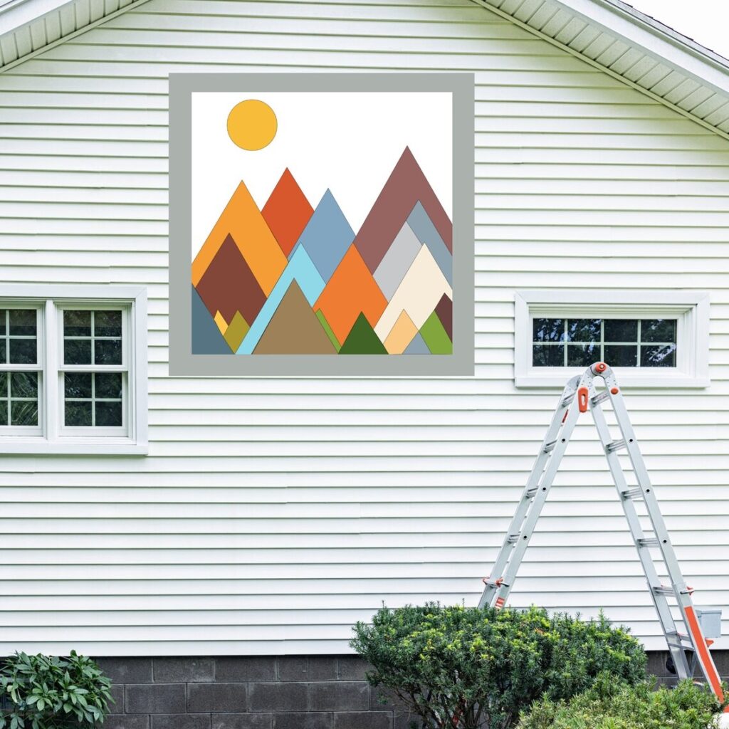 the square barn quilt with mountains and sun patterns hanging on the white barn.