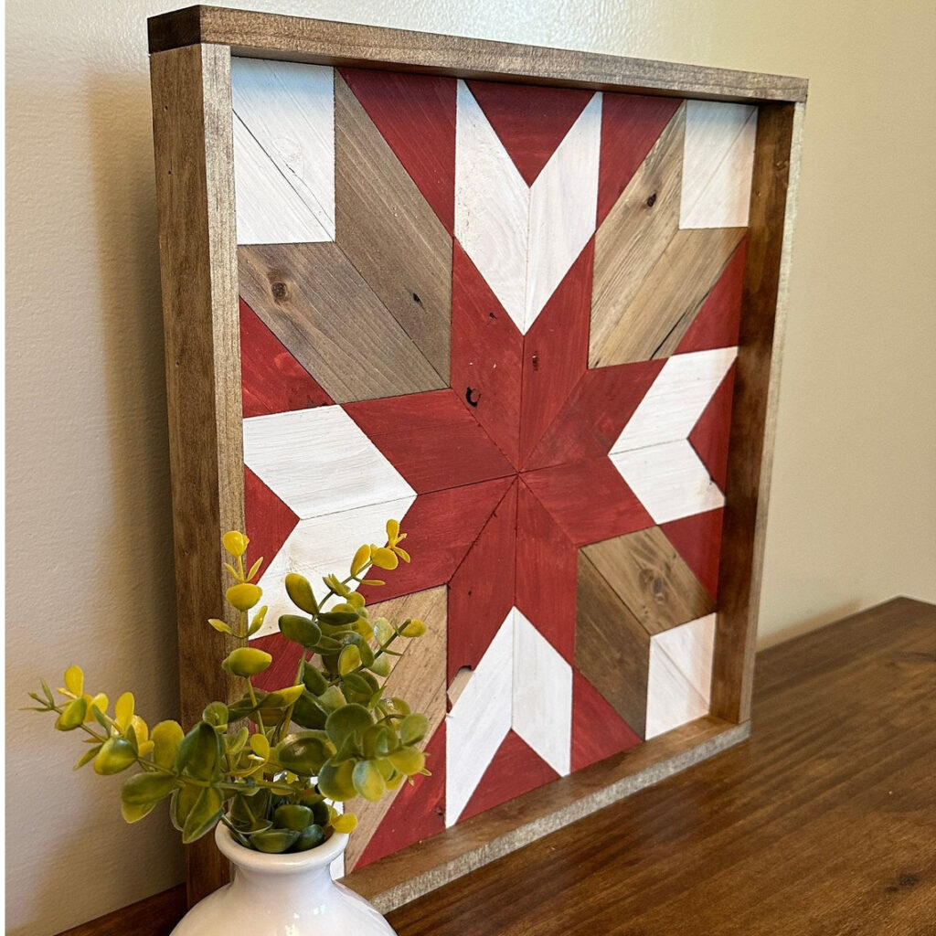 The square barn quilt with red and white star patterns was placed on the wood floor and next to a flower pot.